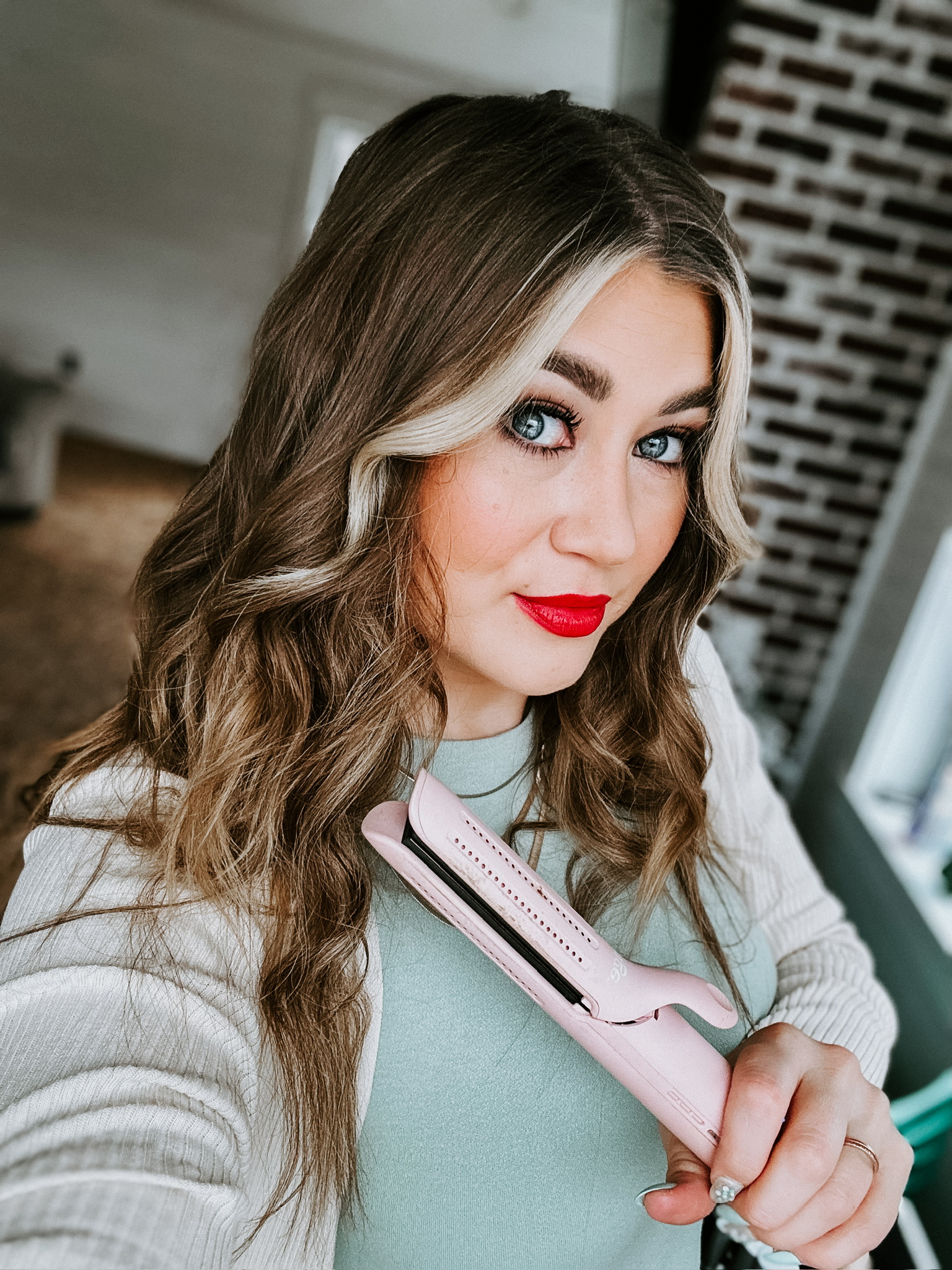 Savannah Kreger styling her long wavy hair with a pink L’ange hair straightener while showcasing smooth curls and vibrant red lipstick in a bright indoor setting