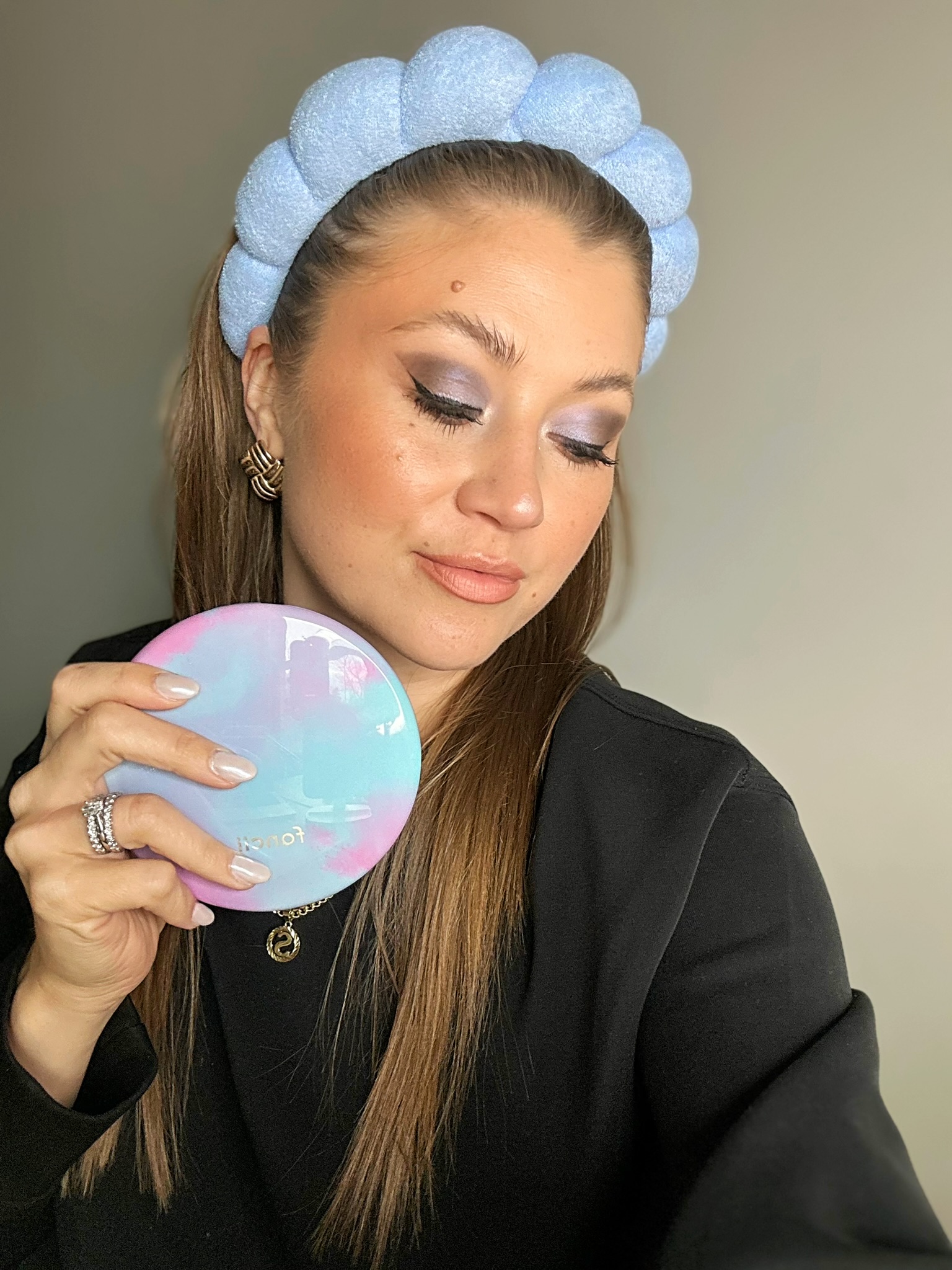 thesavannahk beauty blogger wearing pastel blue padded headband with soft glam makeup and winged eyeliner, holding colorful compact mirror