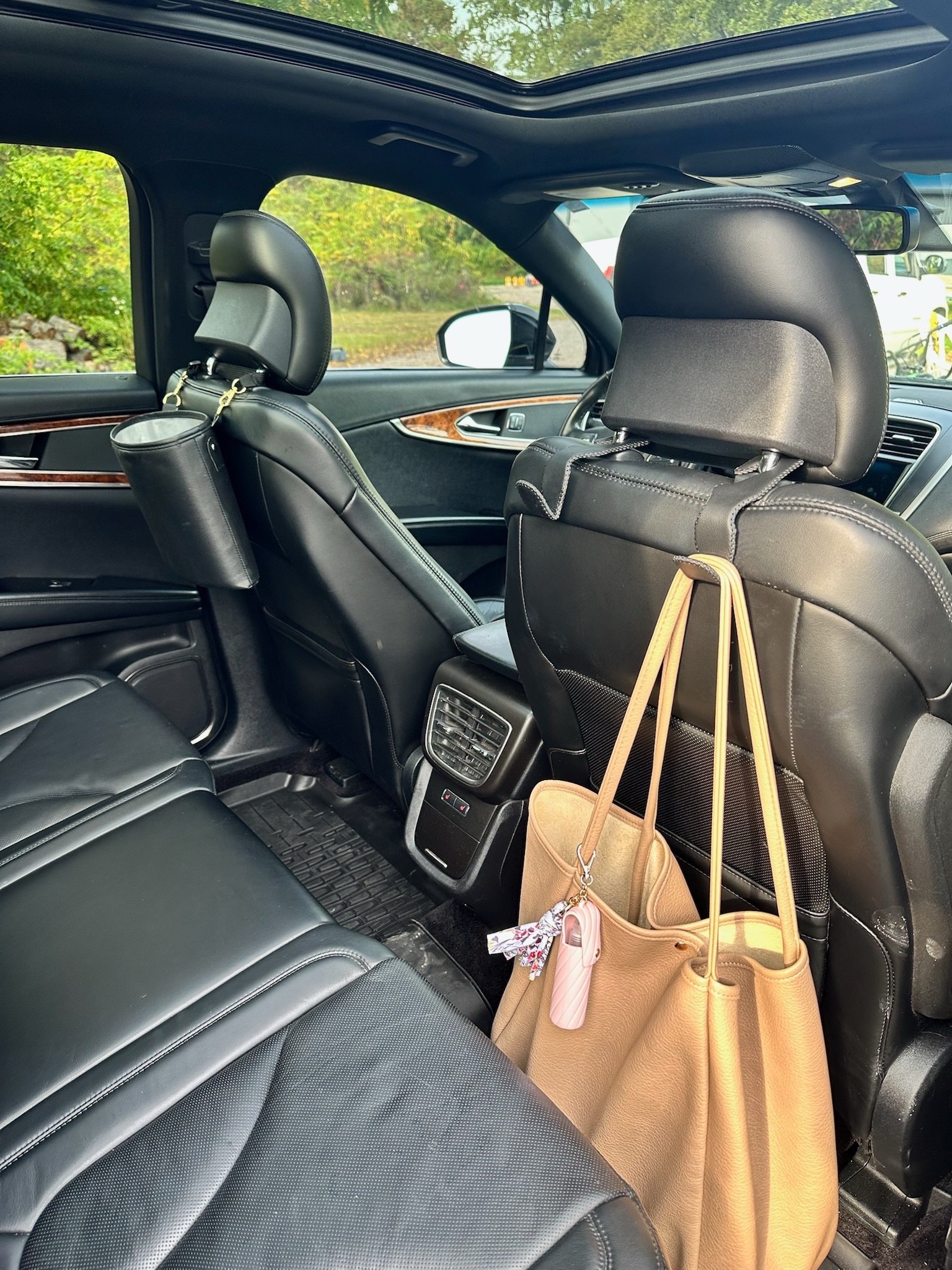thesavannahk car organization hack showing tan tote bag hanging on front seat hook with keychain and pink sanitizer case, black leather interior and family travel accessory setup