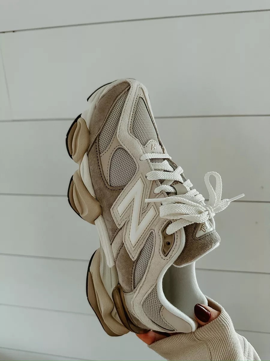A close up photo of beige and cream New Balance 9060 sneakers being held up against a white shiplap wall.