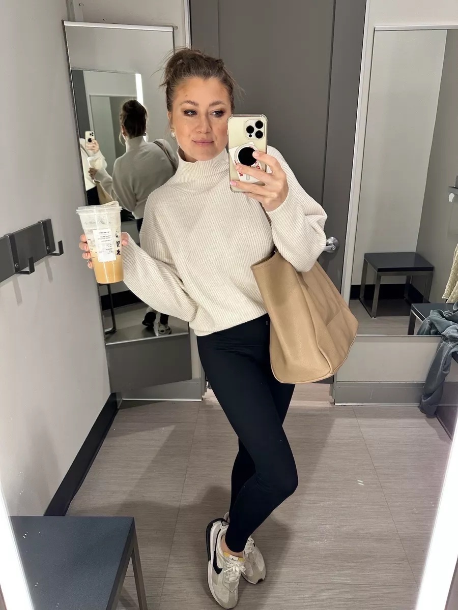 A woman wearing a cream turtleneck sweater, black leggings, and beige New Balance sneakers taking a mirror selfie in a fitting room while holding an iced coffee.