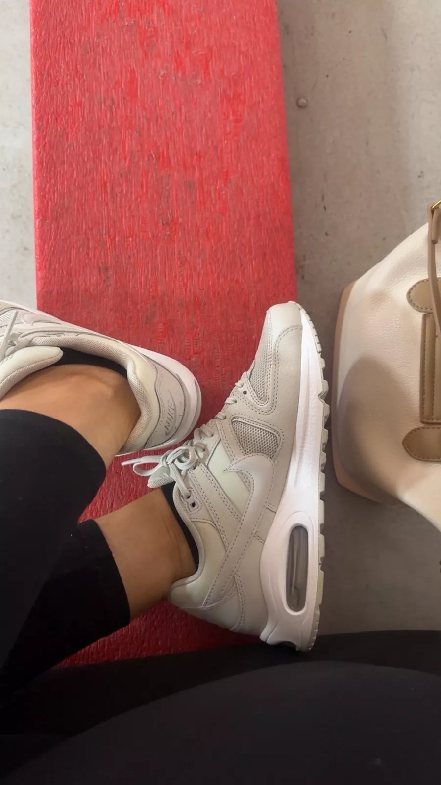A close up of light gray Nike Air Max sneakers paired with black leggings and a beige tote bag resting on a red bench.