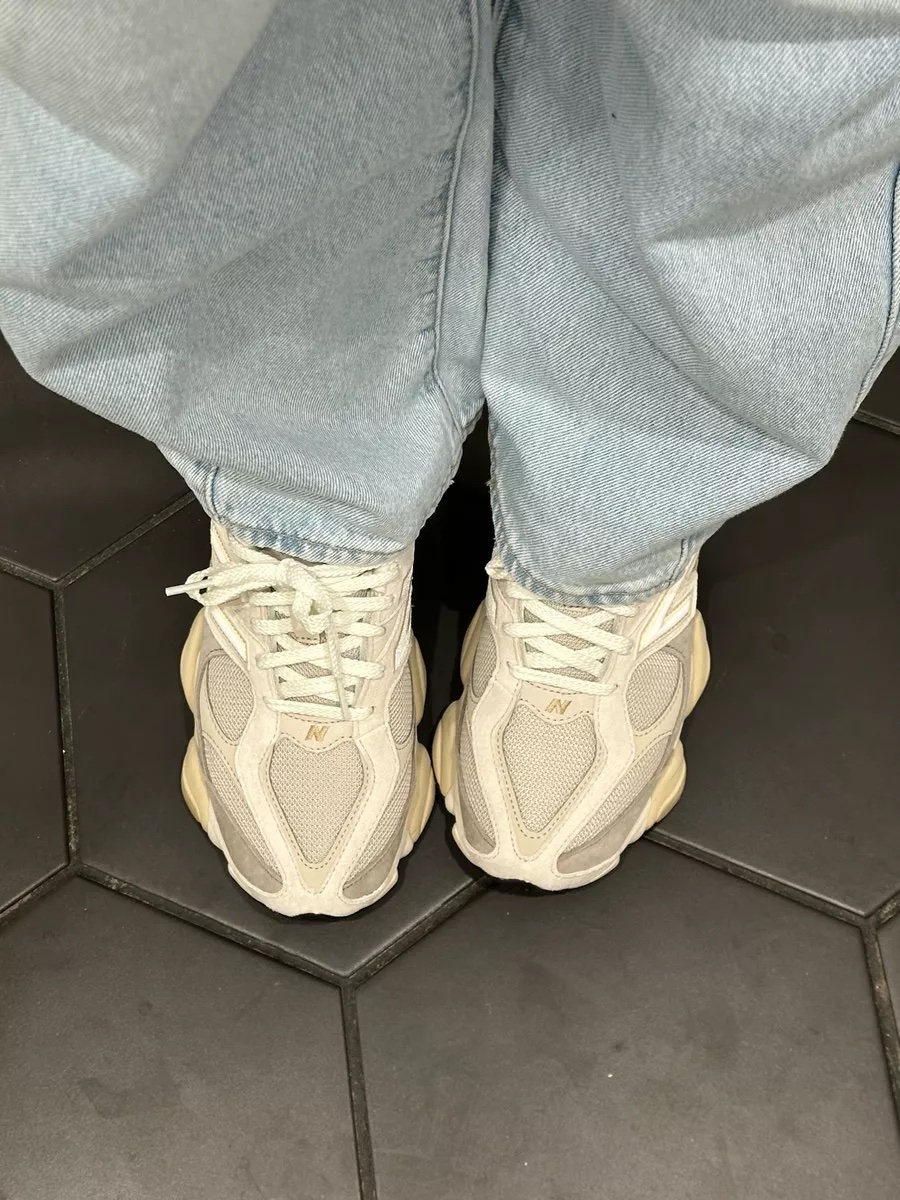 A close up of beige and cream New Balance 9060 sneakers worn with relaxed fit light wash jeans on a black tile floor.