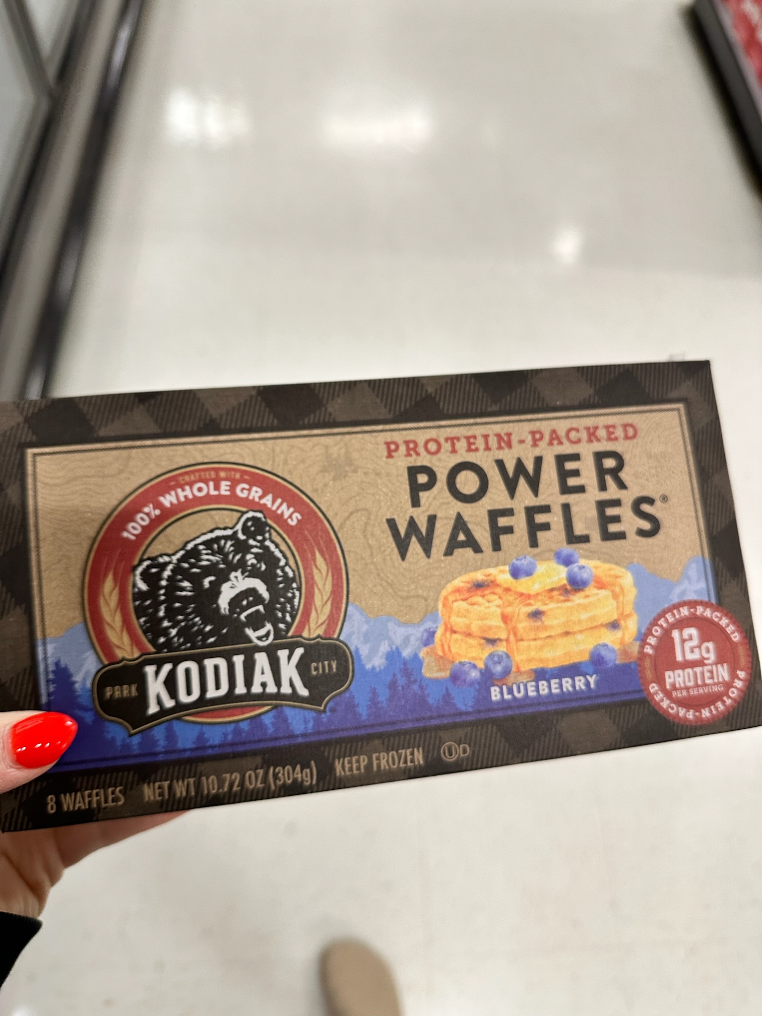 A hand holding a box of Kodiak protein packed blueberry waffles while shopping in a freezer section.
