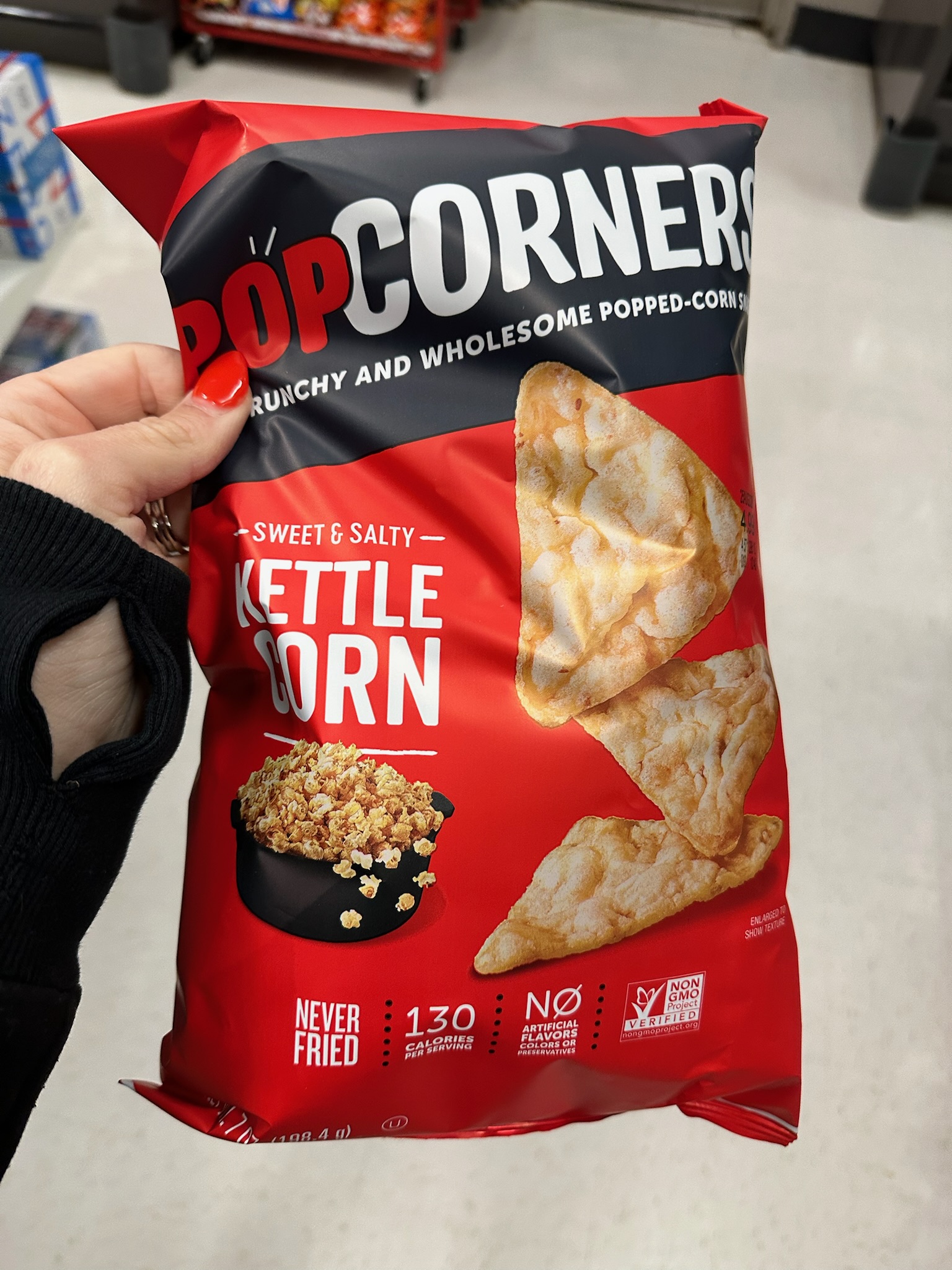 A hand holding a red bag of PopCorners kettle corn chips in a grocery store aisle.
