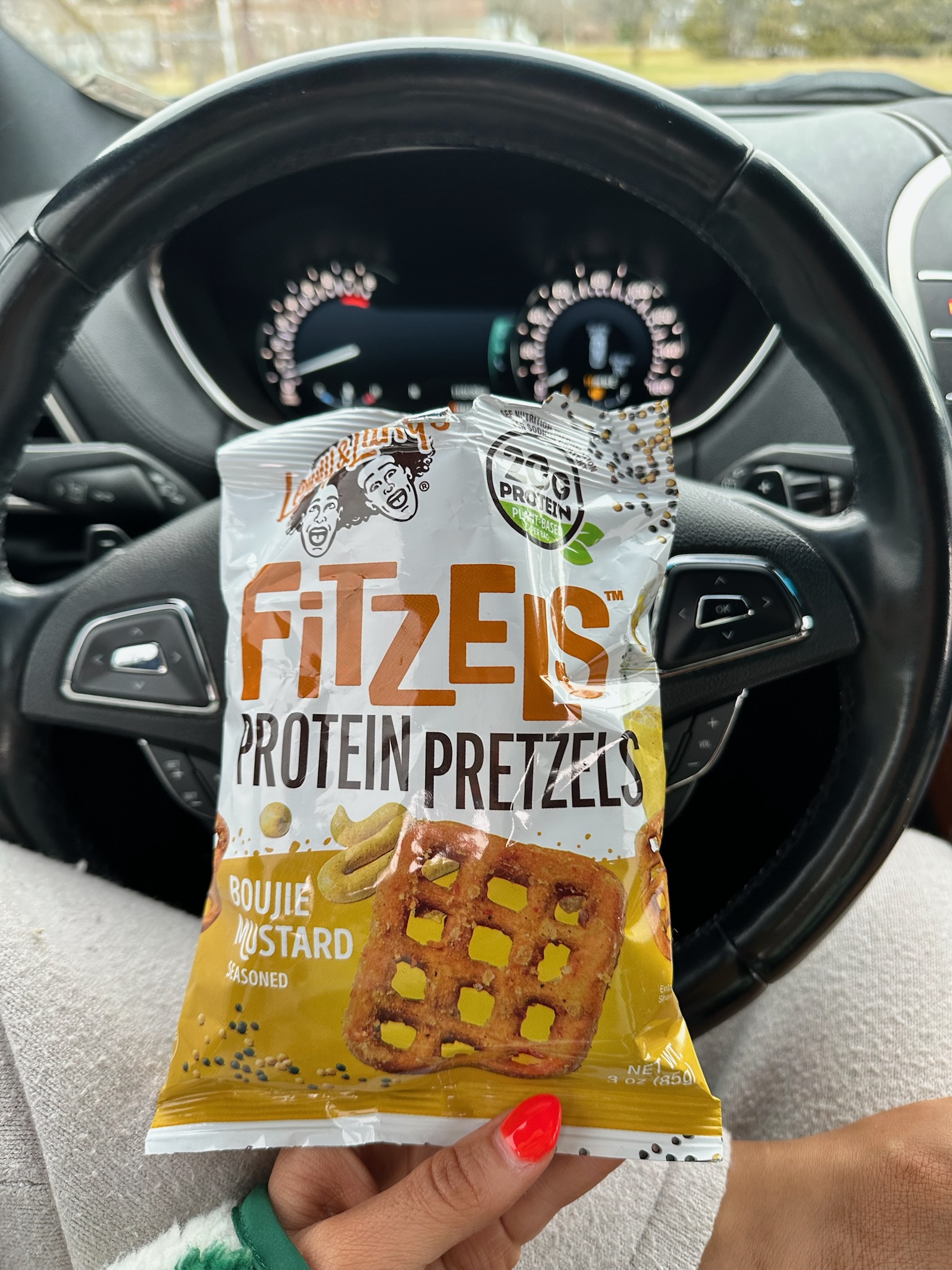 A hand holding a bag of FitZels boujie mustard protein pretzels inside a car with the steering wheel visible.