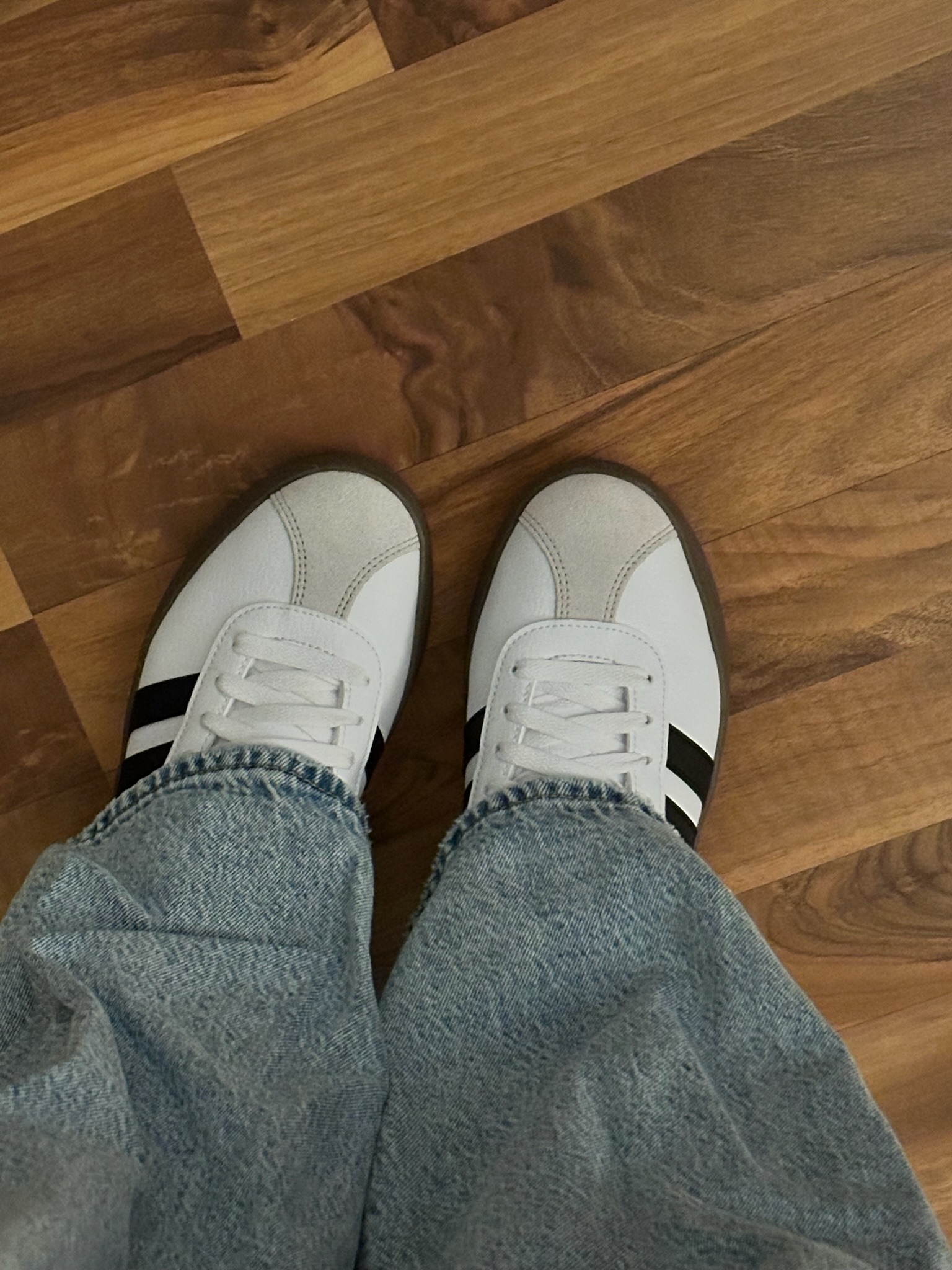 A pair of white Adidas sneakers with black stripes worn with light wash jeans standing on a wood floor.