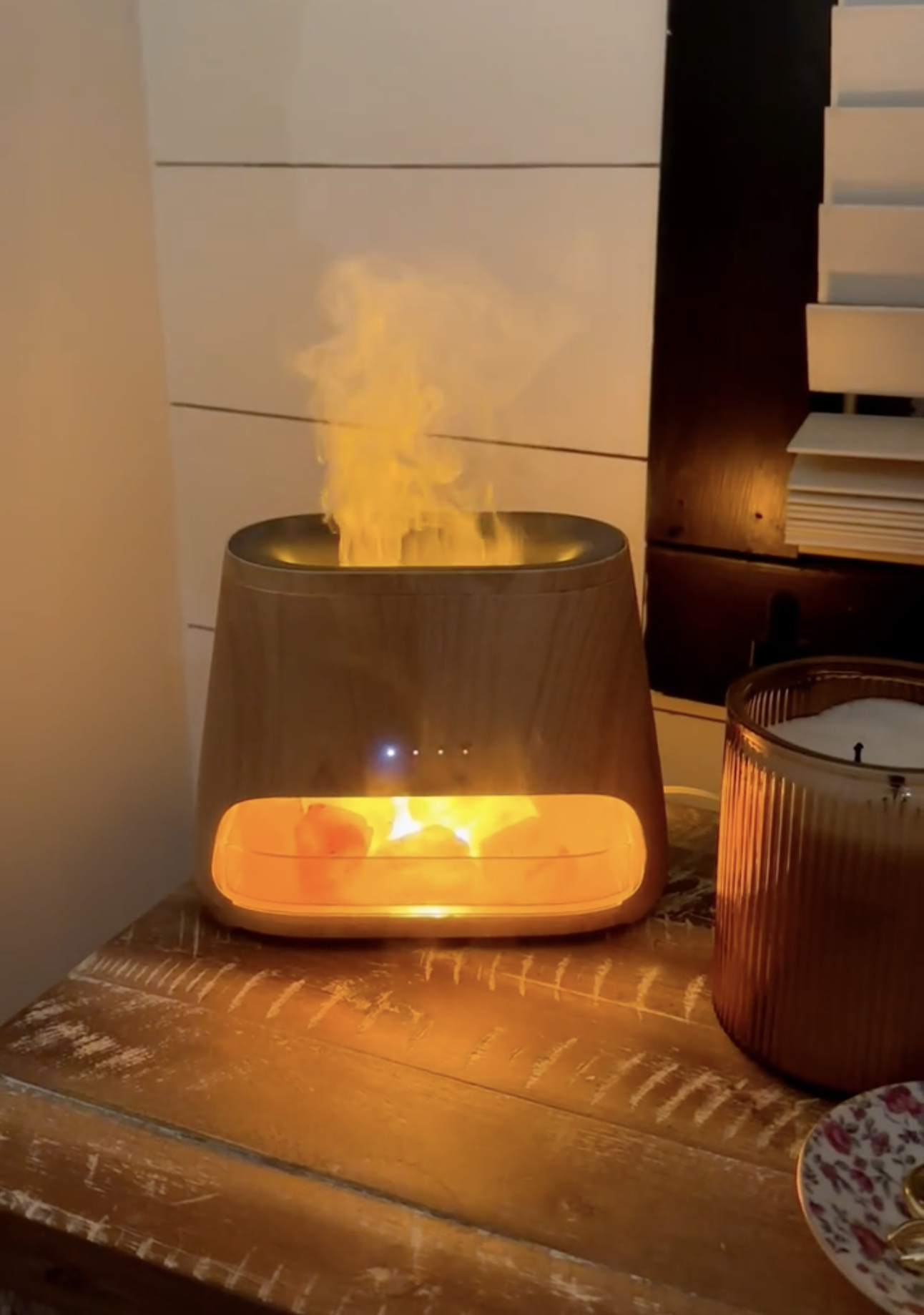 A wooden electric diffuser on a bedside table emitting warm orange light and vapor next to a candle and small dish.
