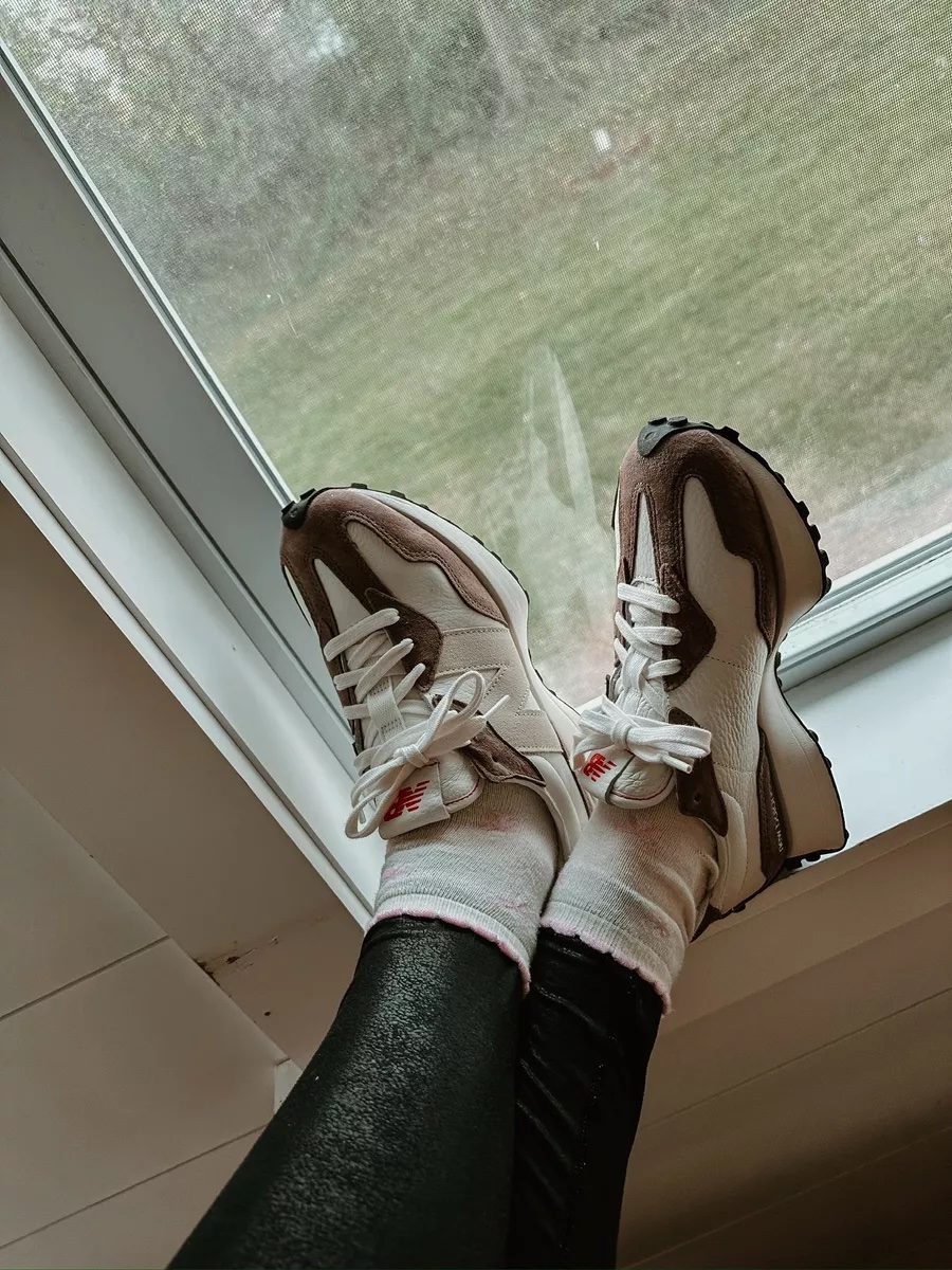 A pair of taupe and white New Balance sneakers worn with white socks and black leggings, propped up near a window.