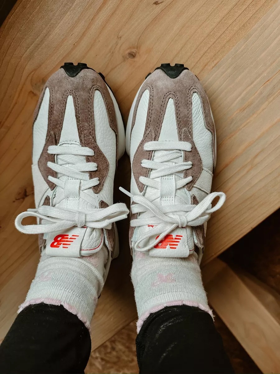A close up of white and taupe New Balance sneakers worn with white socks and black leggings on a wooden floor.