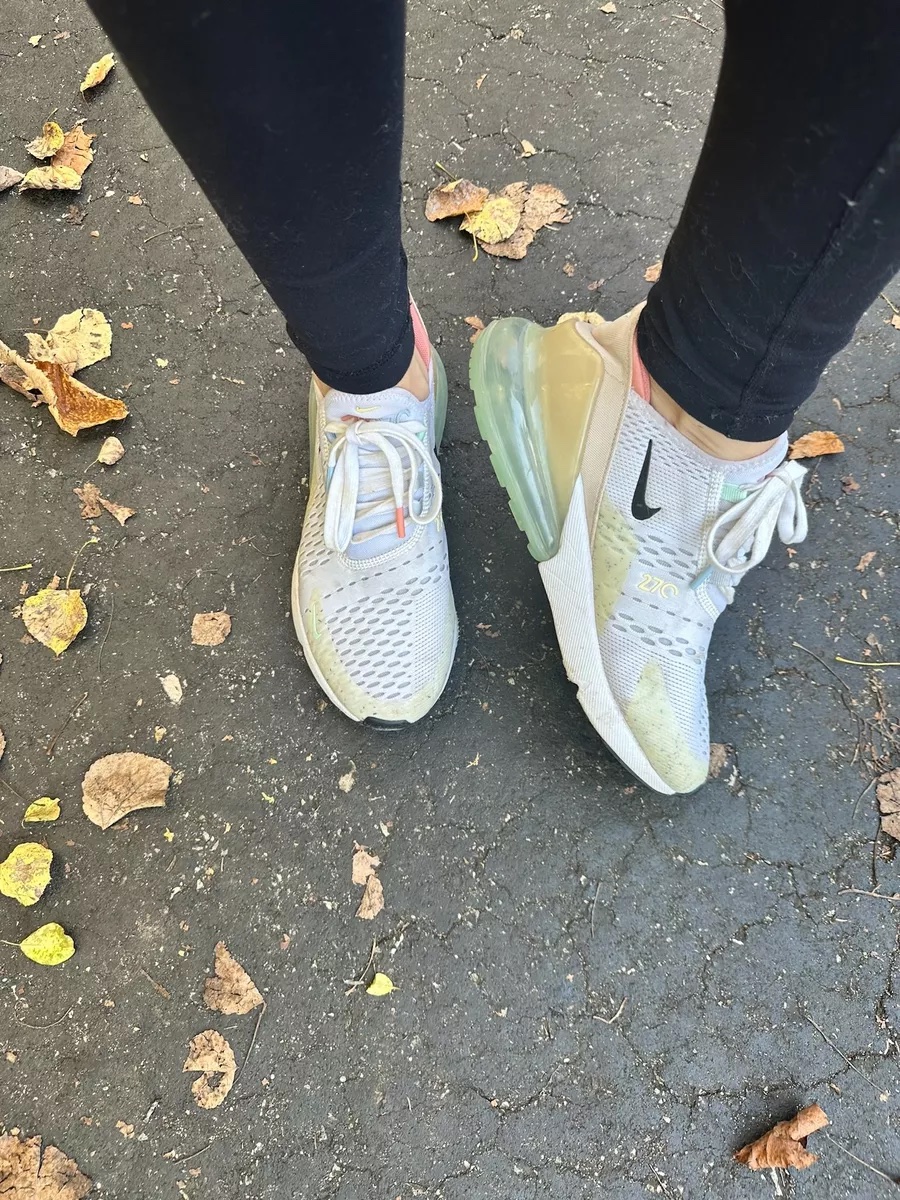 A close up of dusty white Nike Air Max 270 sneakers worn with black leggings while standing on a leaf covered pavement.