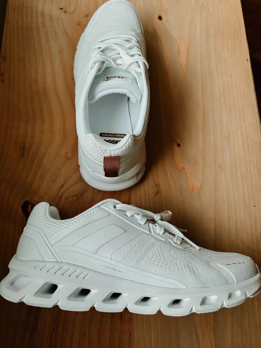 A pair of all white athletic sneakers with a thick, ventilated sole displayed on a wooden surface.